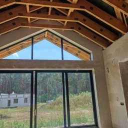 Pogromcy Tynków - Surowy stan wnętrza z drewnianą konstrukcją dachu i dużymi oknami z widokiem na las. Ściany w trakcie prac wykończeniowych, widoczne ślady po szpachlowaniu.