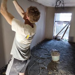 Pogromcy Tynków - Pracownik w trakcie szpachlowania sufitu w pomieszczeniu z widocznymi przewodami elektrycznymi i elementami przygotowania do dalszych prac wykończeniowych.