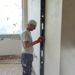 Pogromcy Tynków - Tynkarz w czerwonych rękawicach, z poziomicą, wyrównuje świeżo nałożony tynk na ścianie w jasnym wnętrzu. Widoczne narzędzia i resztki materiałów na podłodze.