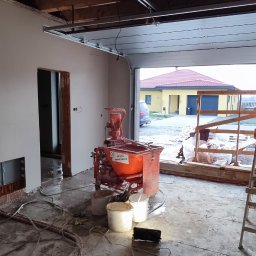 Pogromcy Tynków - Wnętrze w trakcie prac: ściany po tynkowaniu, widoczna maszyna do tynków, kable i wiaderka. W tle brama garażowa i budynek. Surowe drewniane wykończenie sufitu.