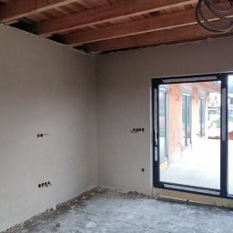 Pogromcy Tynków - Surowy stan wnętrza z drewnianym stropem i ścianami po tynkowaniu. Widoczne puszki elektryczne, okno balkonowe i materiały budowlane na podłodze.