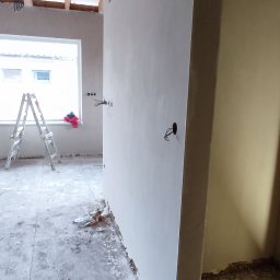 Pogromcy Tynków - Świeżo otynkowana ściana w surowym wnętrzu z drewnianym stropem i aluminiową drabiną. Widoczne kable i gniazdka elektryczne, stan surowy.