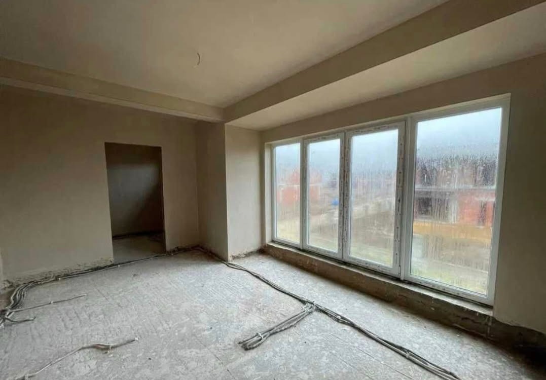 Surowe wnętrze pokoju przed wykończeniem: widoczne kable na podłodze, ściany przygotowane do malowania, duże okno z widokiem na zewnątrz. Etap prac wykończeniowych.