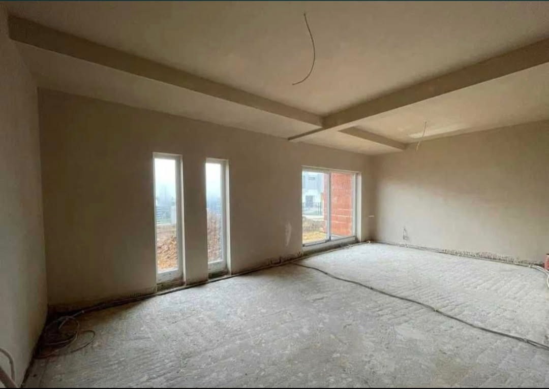 Wnętrze w trakcie prac wykończeniowych: ściany pokryte tynkiem, widoczne okna i sufit z elementami dekoracyjnymi. Podłoga przygotowana do dalszych prac.