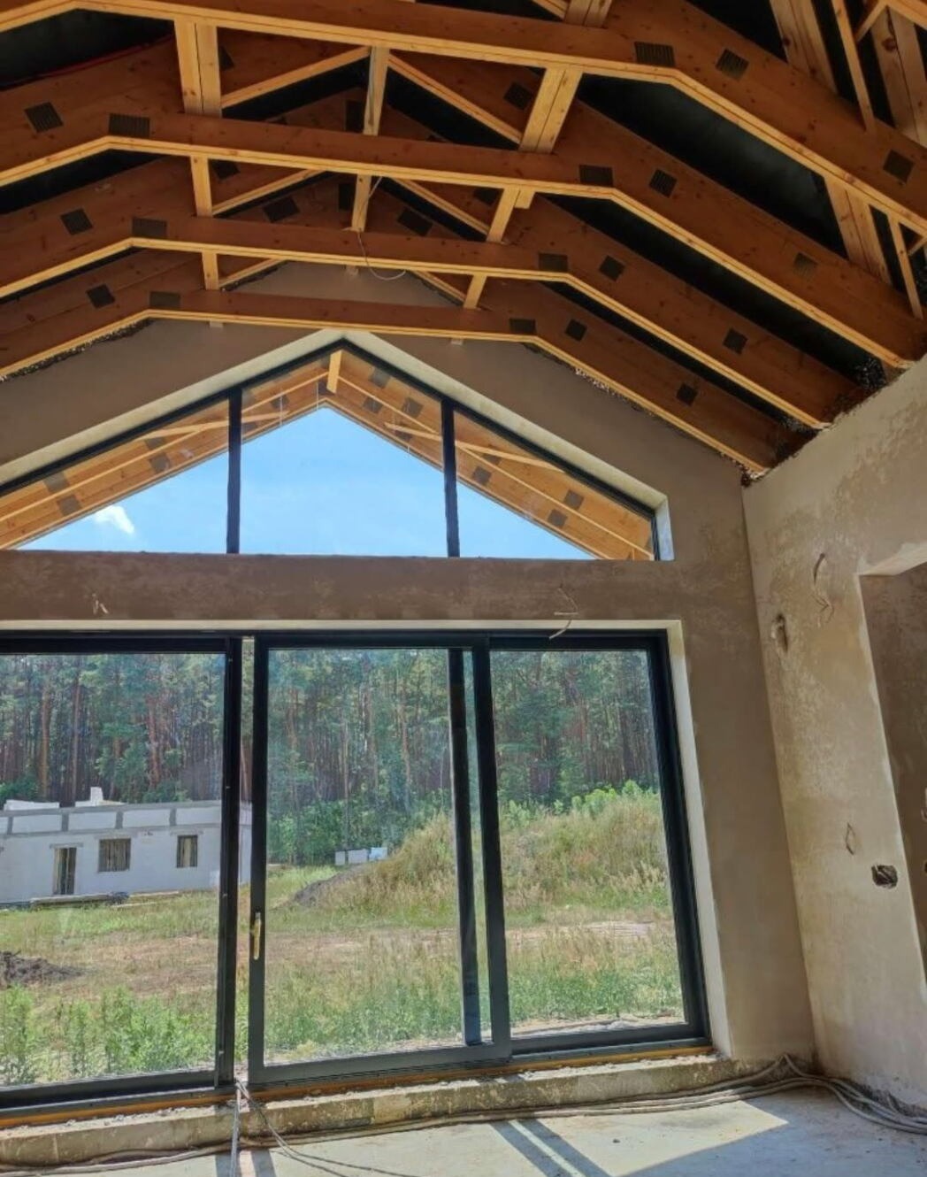 Surowy stan wnętrza z drewnianą konstrukcją dachu i dużym oknem z widokiem na las. Ściany w trakcie prac wykończeniowych, widoczne przewody.