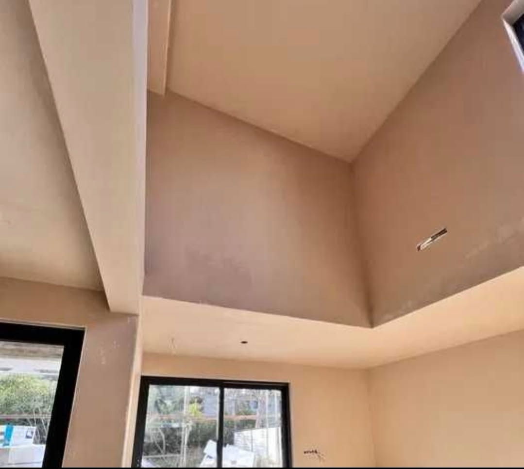 Wnętrze po tynkowaniu: gładkie, beżowe ściany i sufit ze skosem, okna z czarnymi ramami. Widoczne detale wykończenia, świeżo położony tynk.