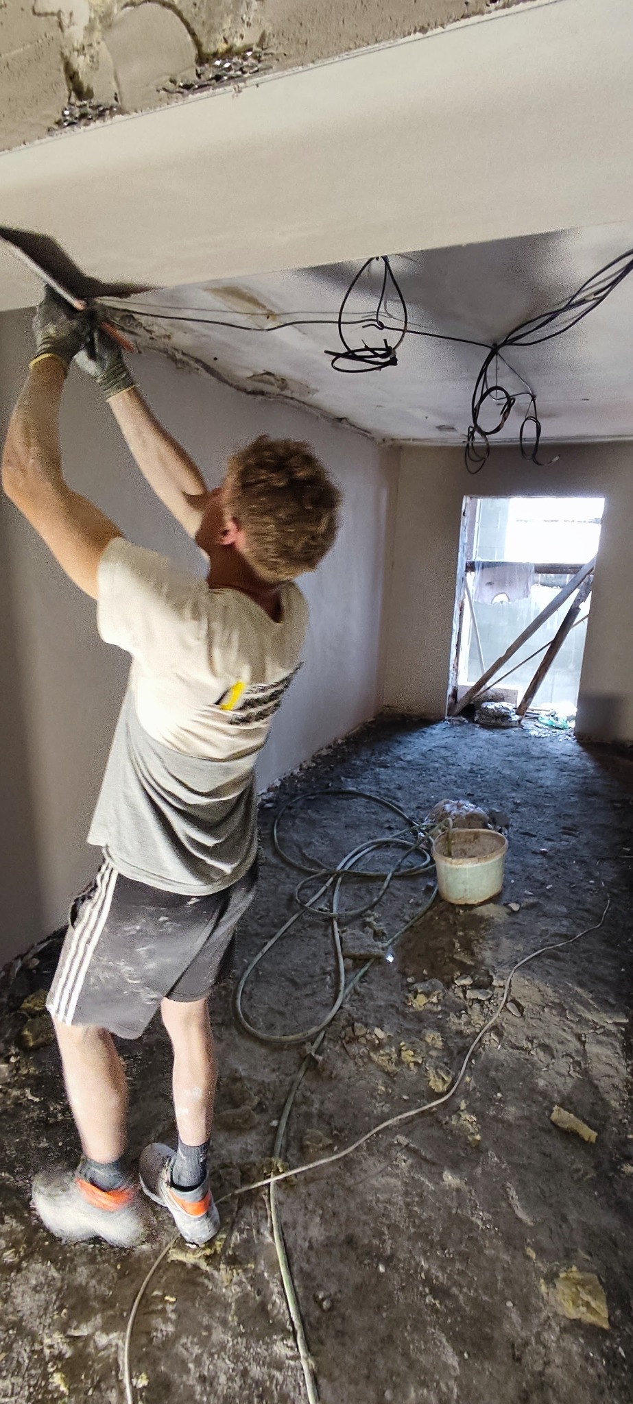Pracownik w trakcie szpachlowania sufitu w pomieszczeniu z widocznymi przewodami elektrycznymi i elementami przygotowania do dalszych prac wykończeniowych.