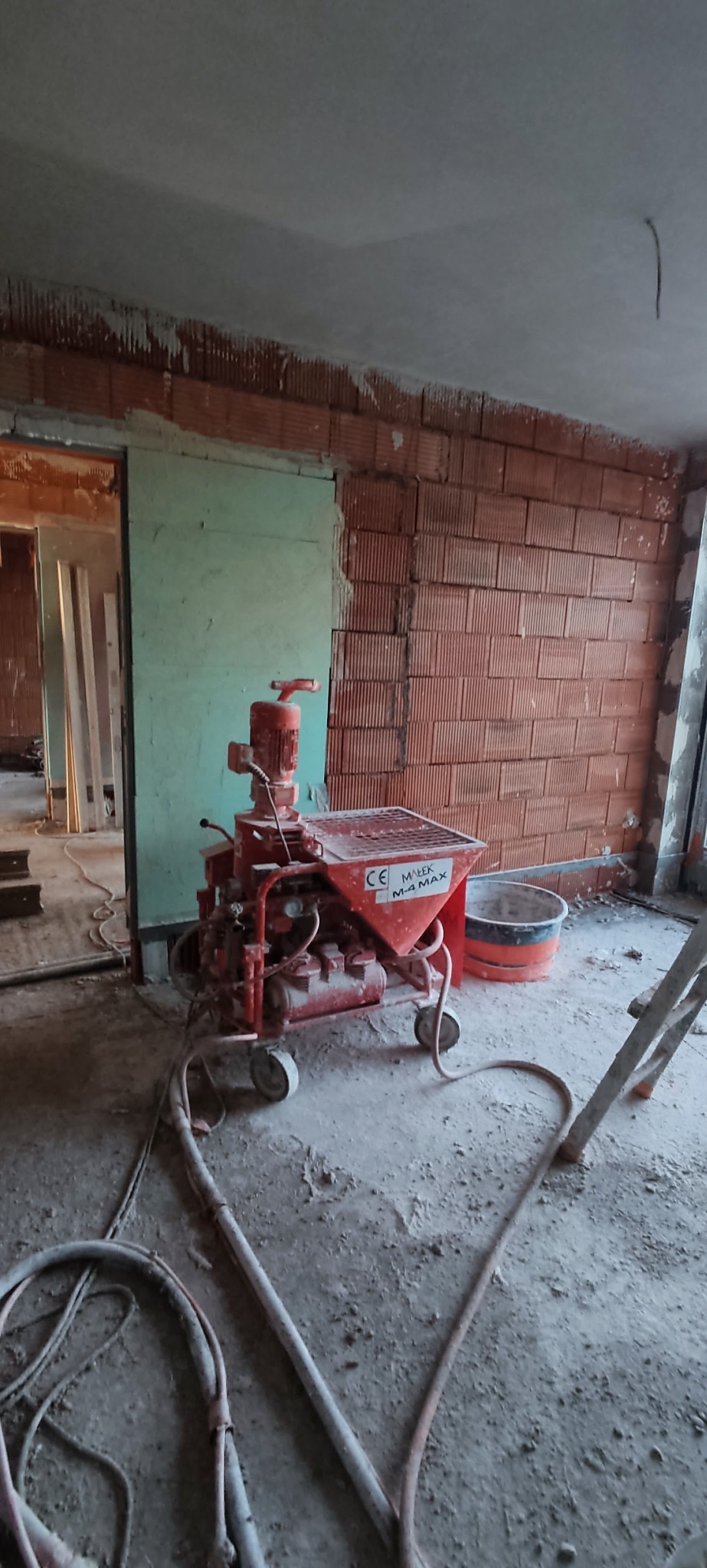 Czerwona maszyna do tynków maszynowych Małek M-4 MAX w surowym wnętrzu z ceglaną ścianą i częściowo otynkowanym wejściem. W tle widoczne przewody i wiadro.