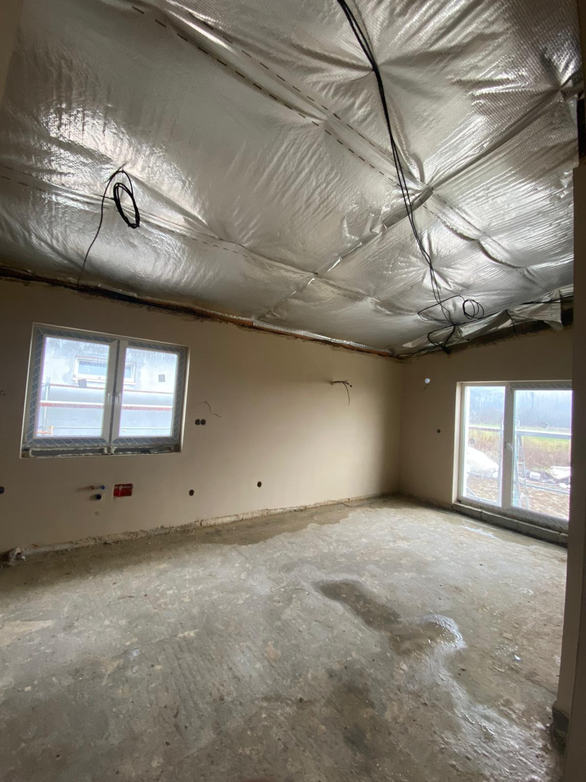 Wnętrze w trakcie prac wykończeniowych: ściany po tynkowaniu, folia na suficie, widoczne instalacje elektryczne, okna PCV. Surowy stan podłogi.