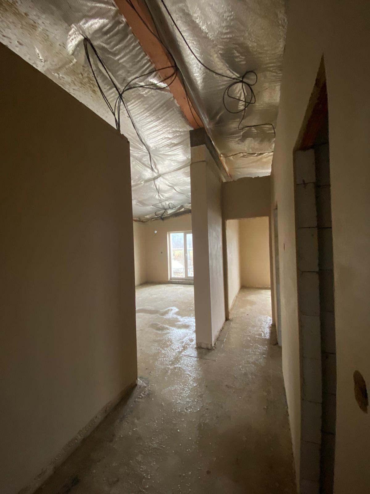 Wnętrze w stanie deweloperskim: surowy beton, ściany po wstępnym wykończeniu, folia na suficie z widocznymi kablami. Widok z korytarza na pokoje.