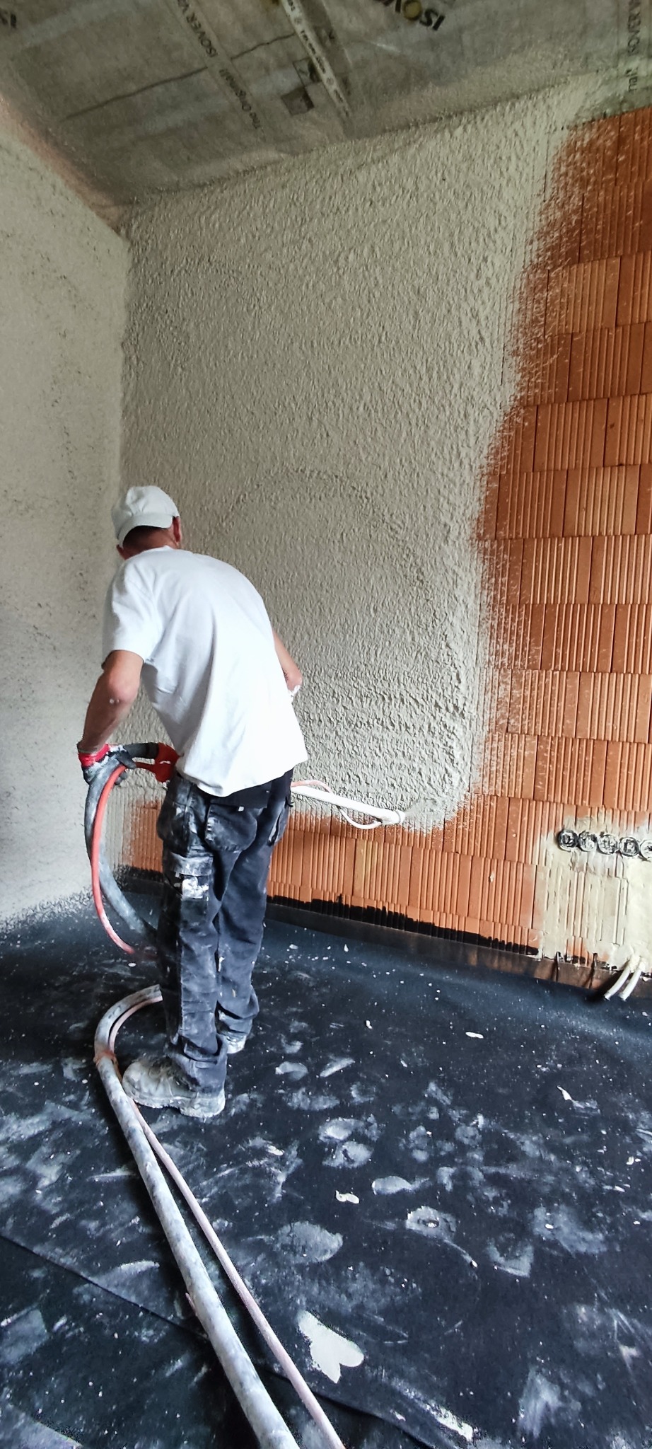 Pracownik w trakcie nakładania tynku maszynowego na ścianę z cegły. Widoczne przewody i narzędzia. Częściowo otynkowana ściana. Praca wewnątrz budynku.