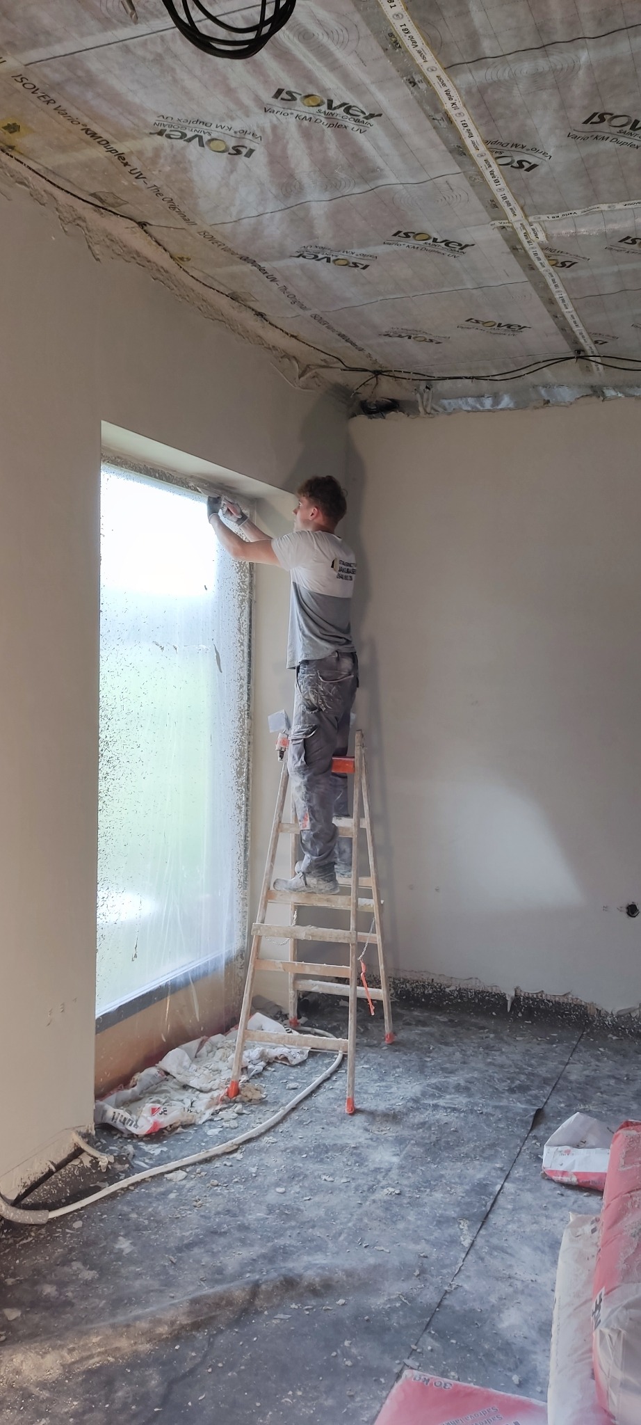 Tynkarz na drabinie szpachluje ścianę przy oknie w pomieszczeniu z folią ochronną na suficie. Widoczne worki z zaprawą i narzędzia.