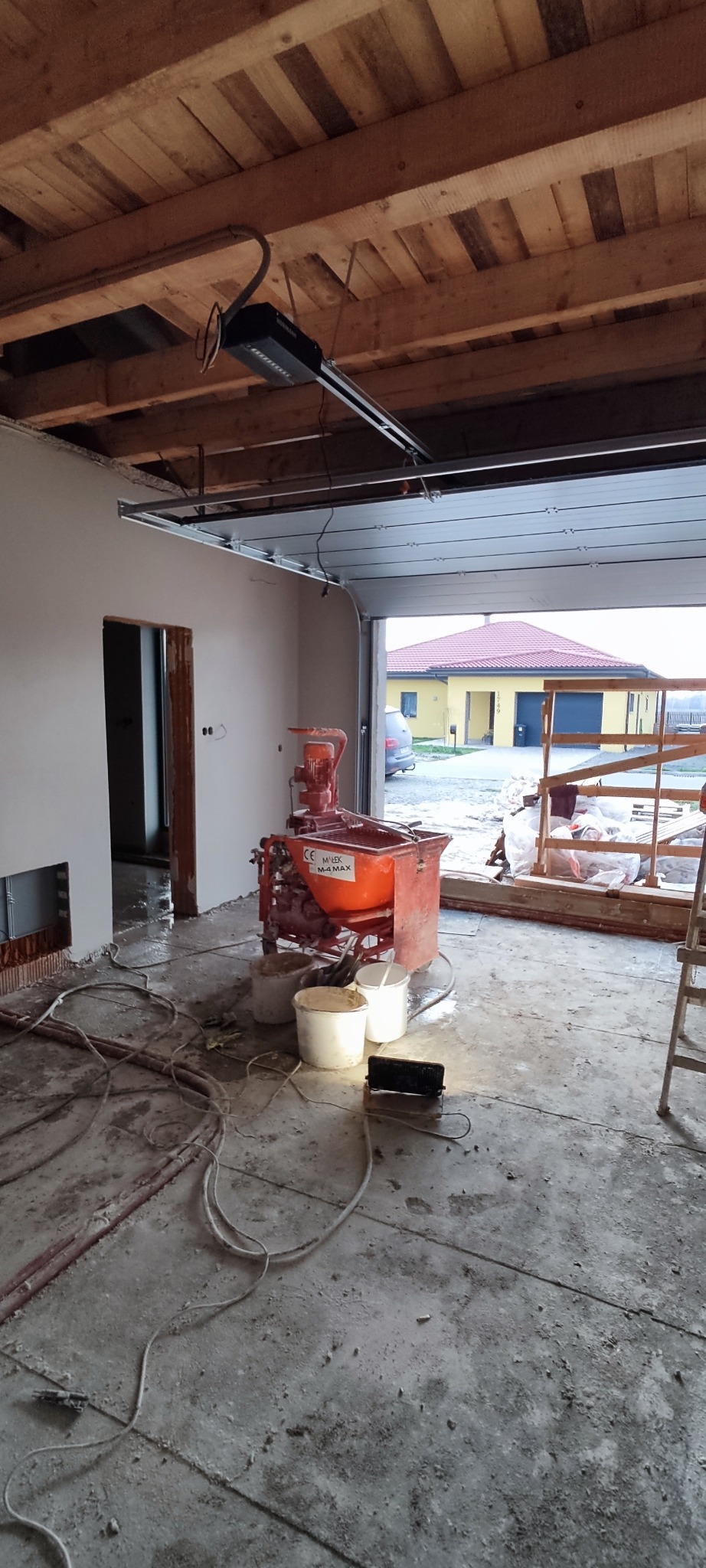 Wnętrze w trakcie prac: ściany po tynkowaniu, widoczna maszyna do tynków, kable i wiaderka. W tle brama garażowa i budynek. Surowe drewniane wykończenie sufitu.