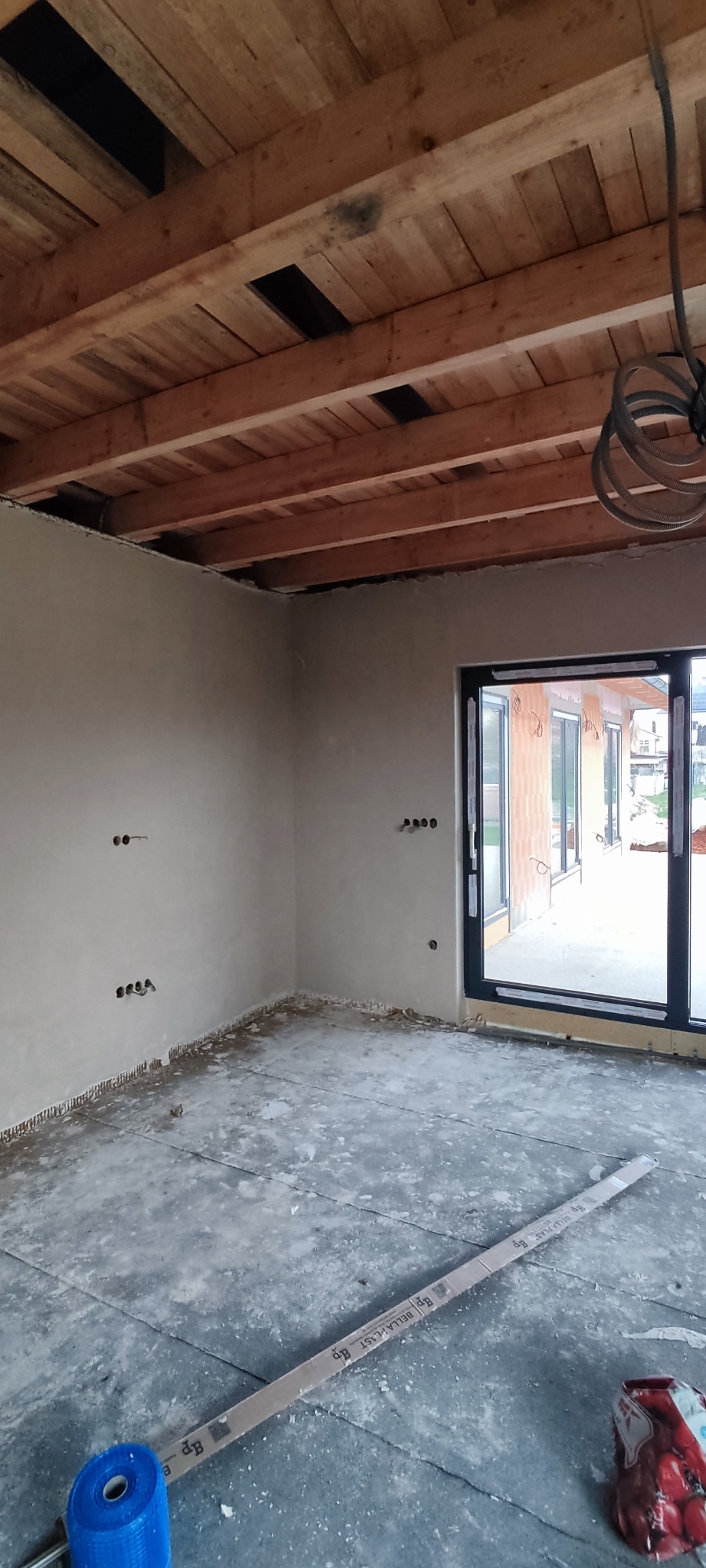 Surowy stan wnętrza z drewnianym stropem i ścianami po tynkowaniu. Widoczne puszki elektryczne, okno balkonowe i materiały budowlane na podłodze.