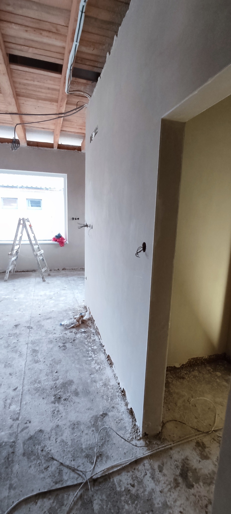 Świeżo otynkowana ściana w surowym wnętrzu z drewnianym stropem i aluminiową drabiną. Widoczne kable i gniazdka elektryczne, stan surowy.