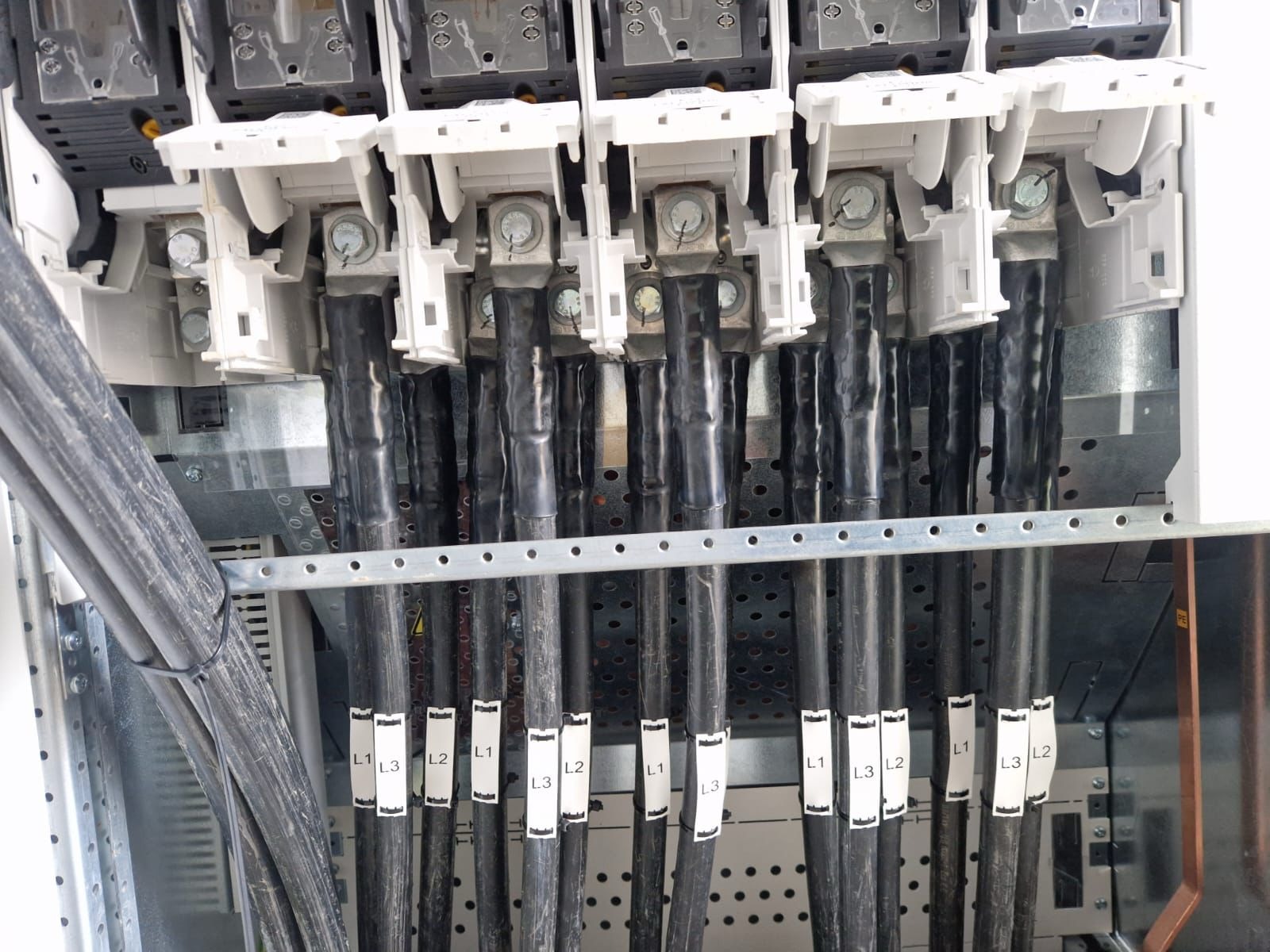 Szczegółowe ujęcie podłączenia przewodów elektrycznych w rozdzielni, z wyraźnie oznaczonymi fazami L1, L2, L3. Widoczne zaciski i metalowa konstrukcja szafy.