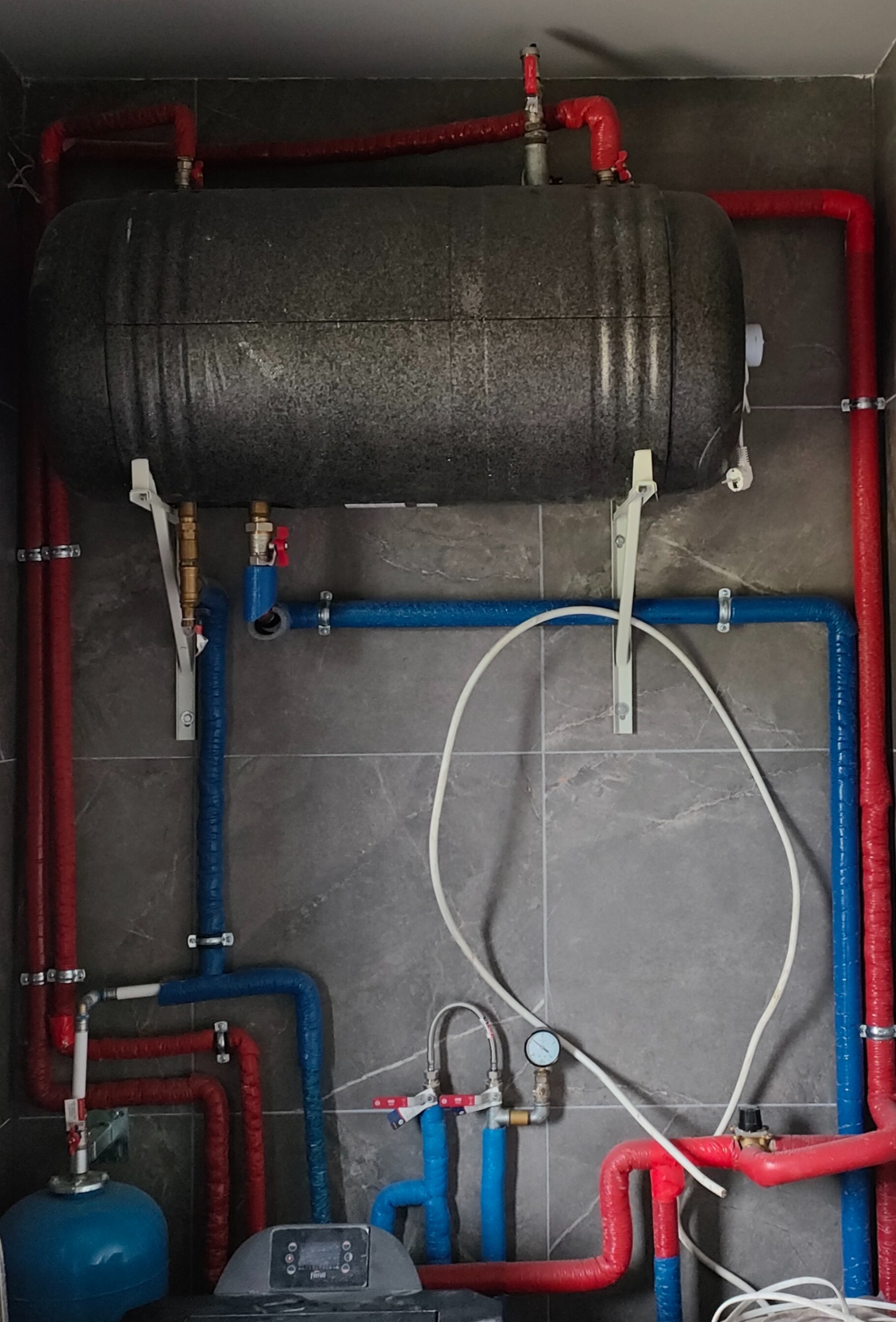 Szczegółowe ujęcie kotłowni z szarymi płytkami. Widoczne: zbiornik, czerwone i niebieskie rury z izolacją, manometr oraz fragment kotła Ferroli. Instalacja po remoncie.