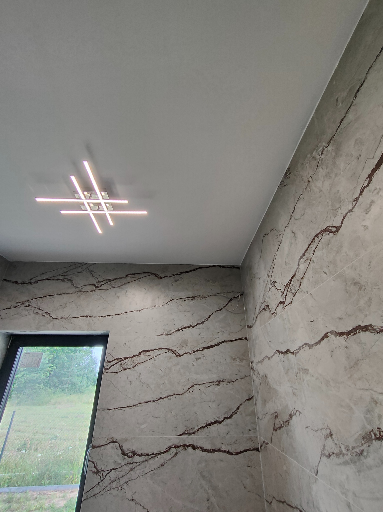 Wykończona łazienka z płytkami imitującymi marmur z brązowymi żyłami, nowoczesna lampa sufitowa, okno z widokiem na zieleń. Jasne, neutralne barwy.