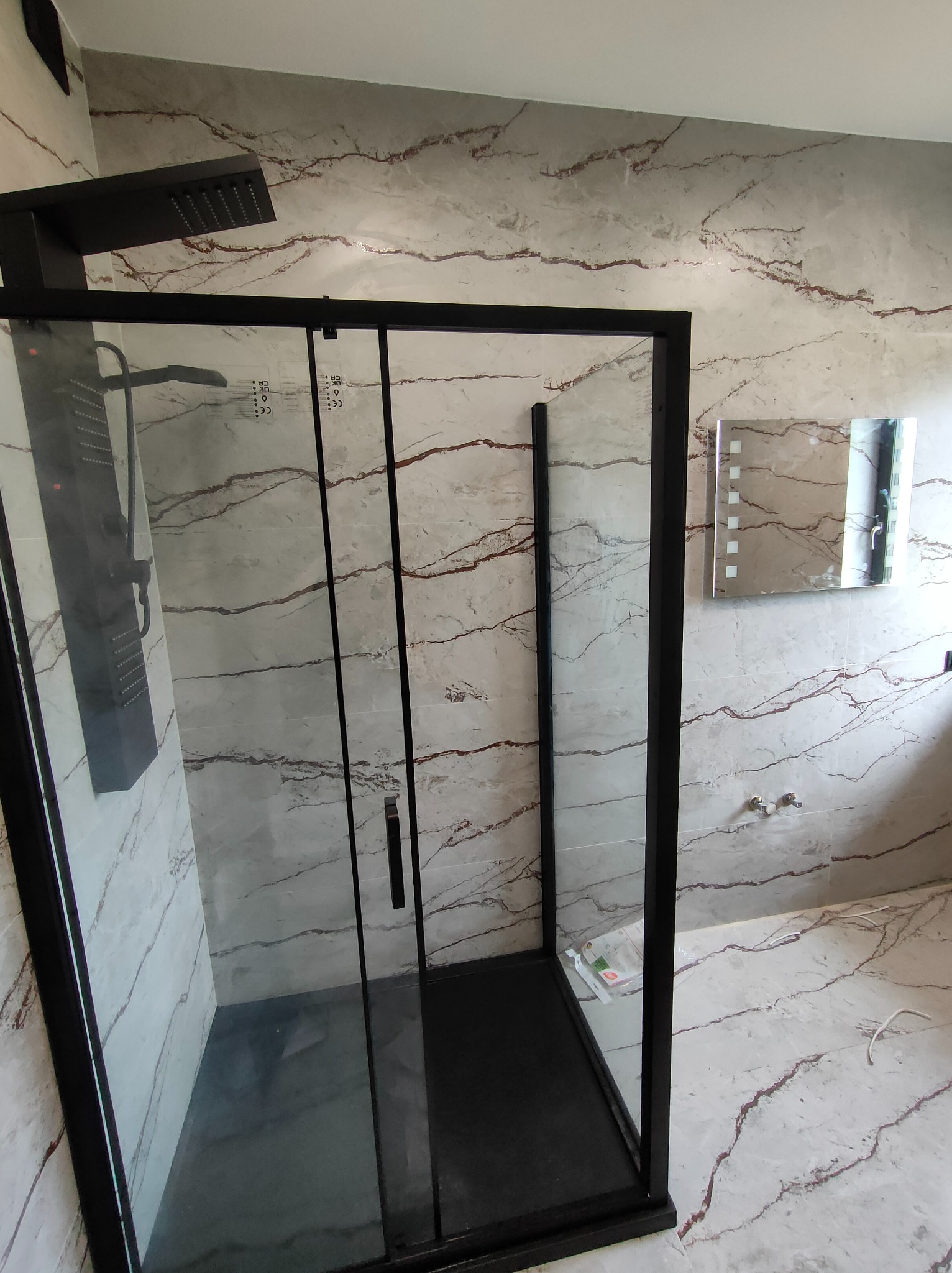 Nowoczesna kabina prysznicowa z czarną armaturą na tle marmurowych płytek w łazience. Widoczne lustro z podświetleniem LED.