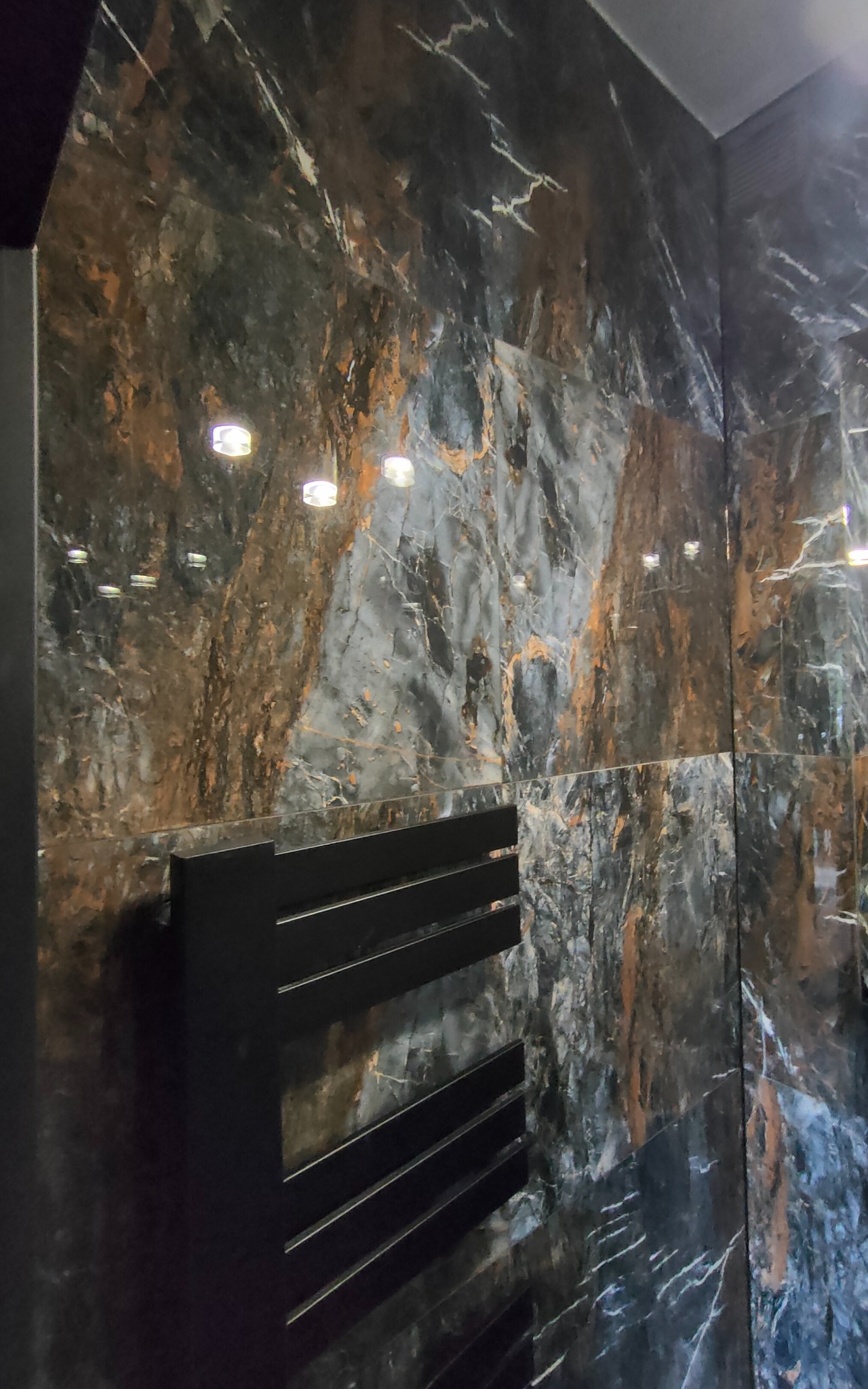 Wykończona ściana w łazience z płytkami imitującymi marmur w odcieniach szarości, brązu i czerni. Widoczny czarny, metalowy grzejnik drabinkowy.