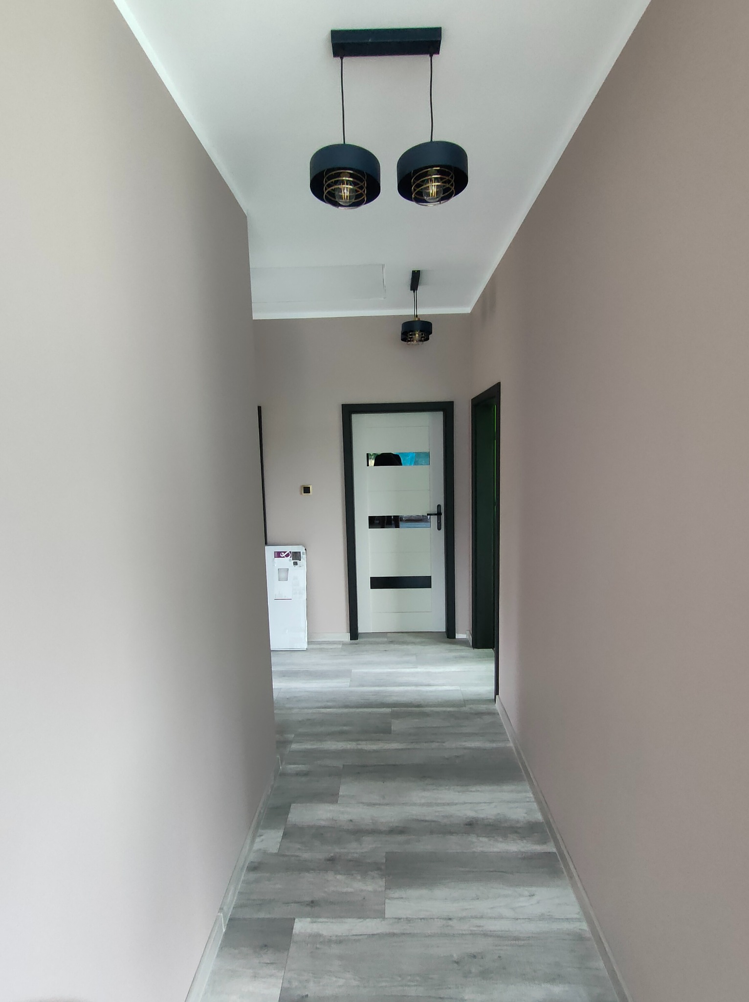 Wykończony korytarz w odcieniach beżu z szarą podłogą imitującą drewno. Widoczne drzwi z przeszkleniami i nowoczesne, czarne lampy sufitowe z dekoracyjnymi żarówkami.