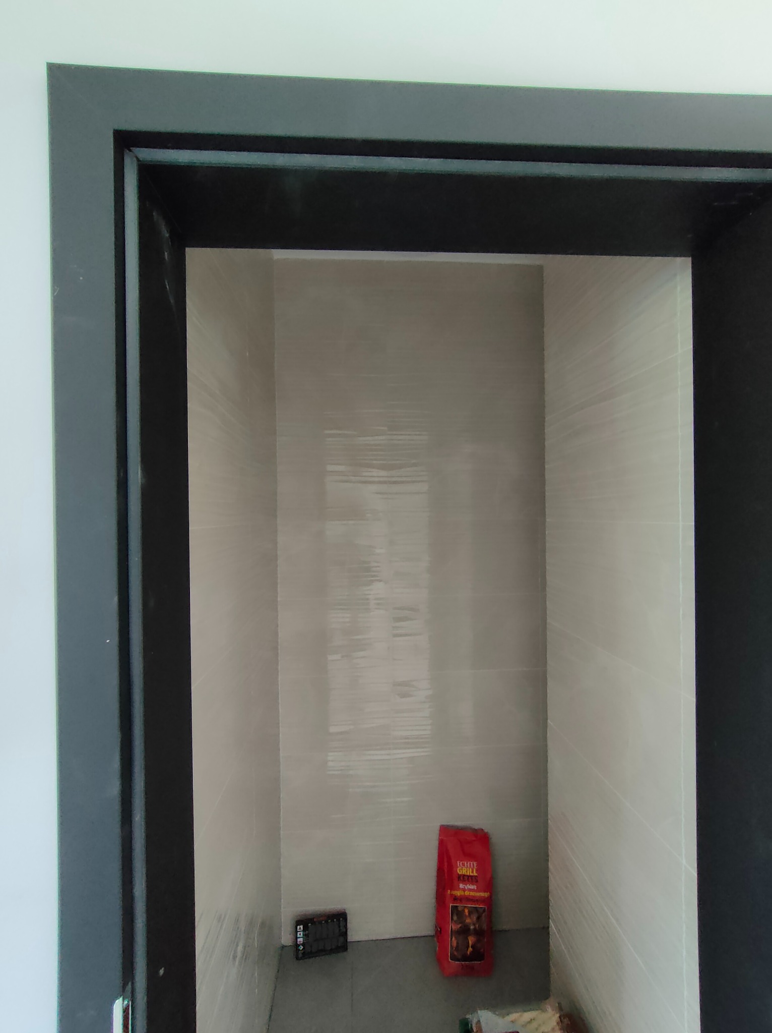 Wykończona wnęka z jasnymi płytkami, widoczna przez czarną futrynę drzwi. Na podłodze torba z brykietem i narzędzia. Minimalistyczny design łazienki.