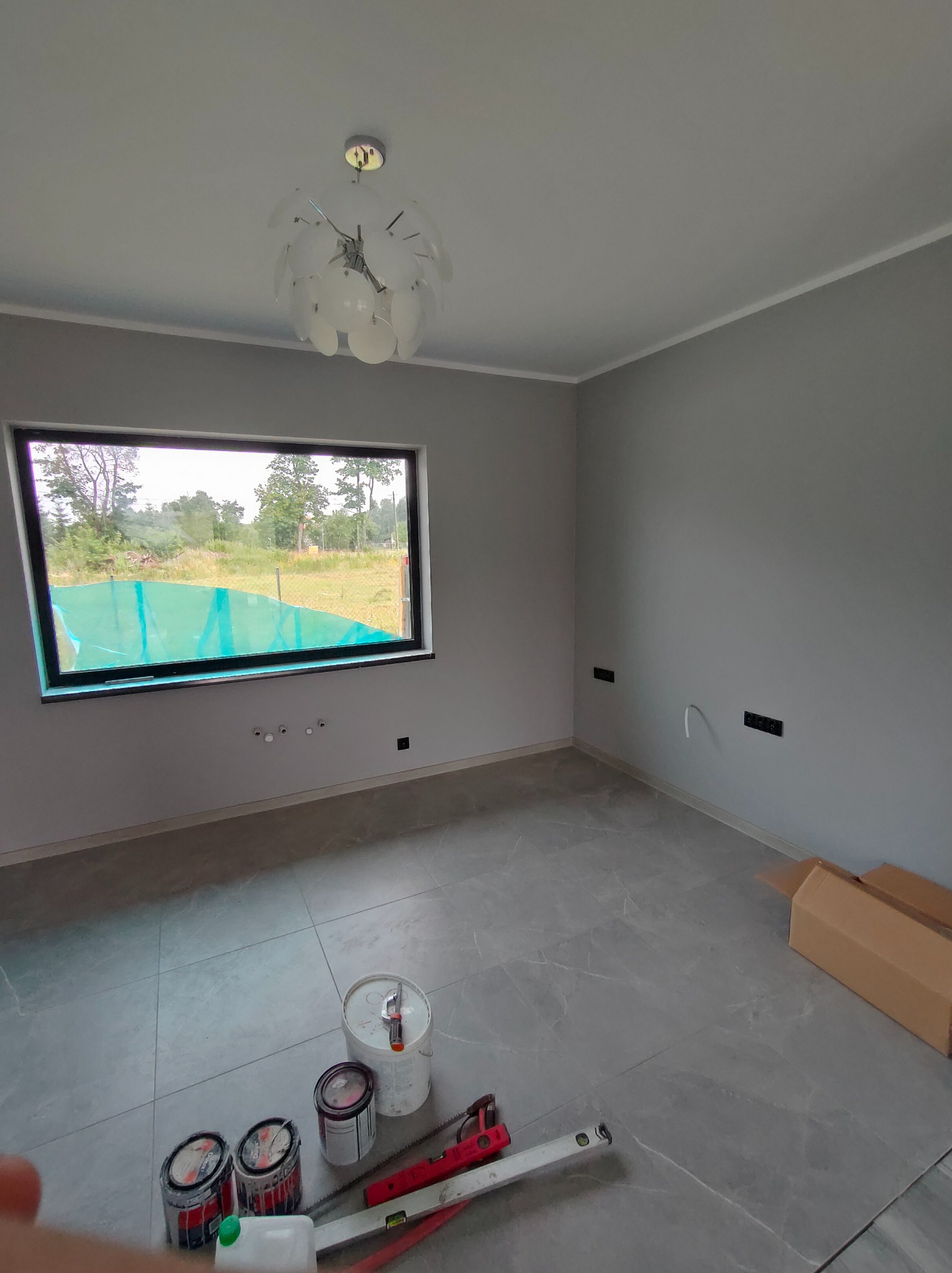 Wnętrze po malowaniu ścian na szaro z widocznymi puszkami farb i narzędziami na podłodze z szarych płytek, okno z widokiem na zieleń.