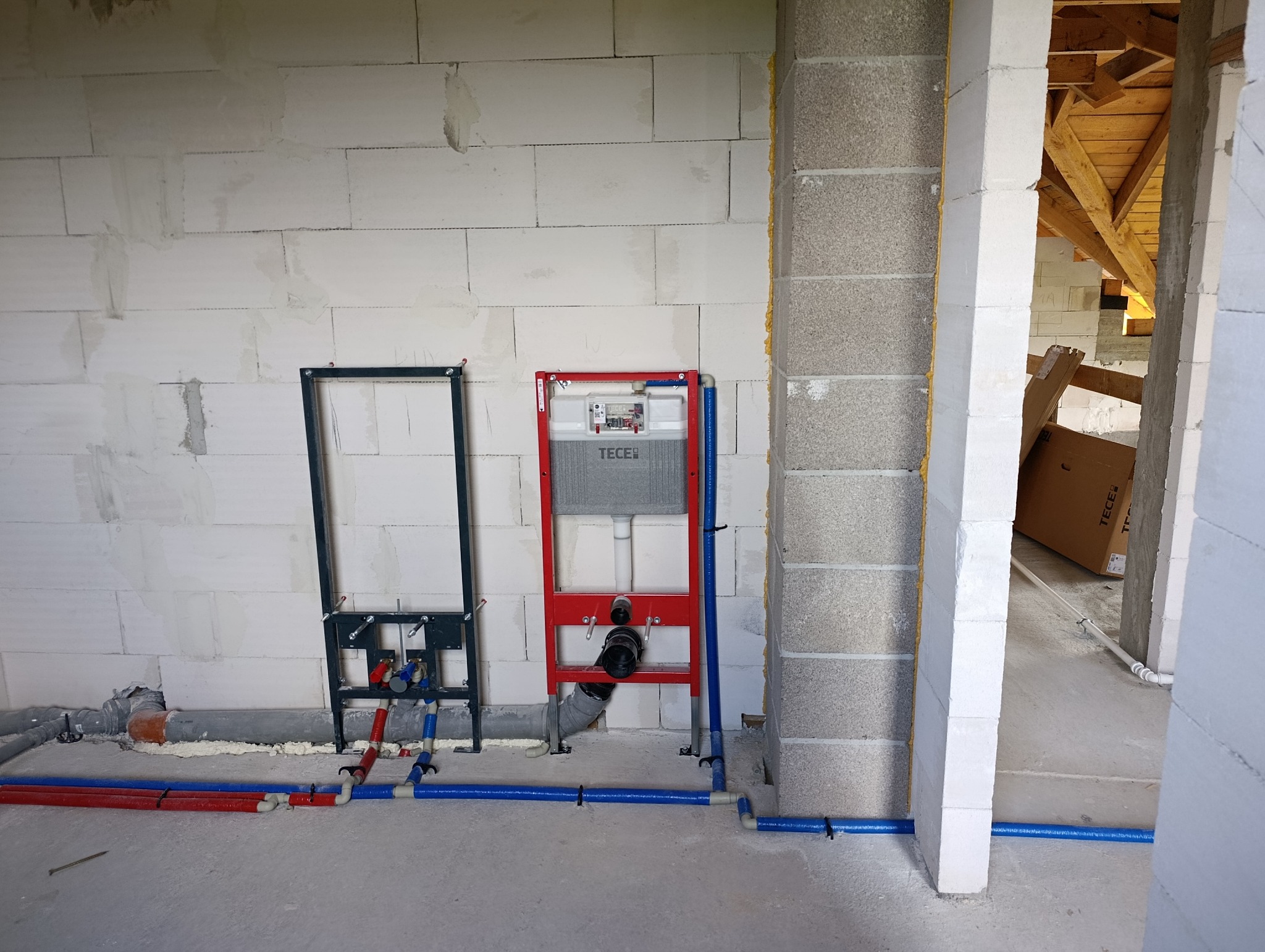 Montaż stelaża podtynkowego TECE i instalacji wodnej w surowym wnętrzu. Widoczne rury PEX w kolorach niebieskim i czerwonym. Ściany z betonu komórkowego.