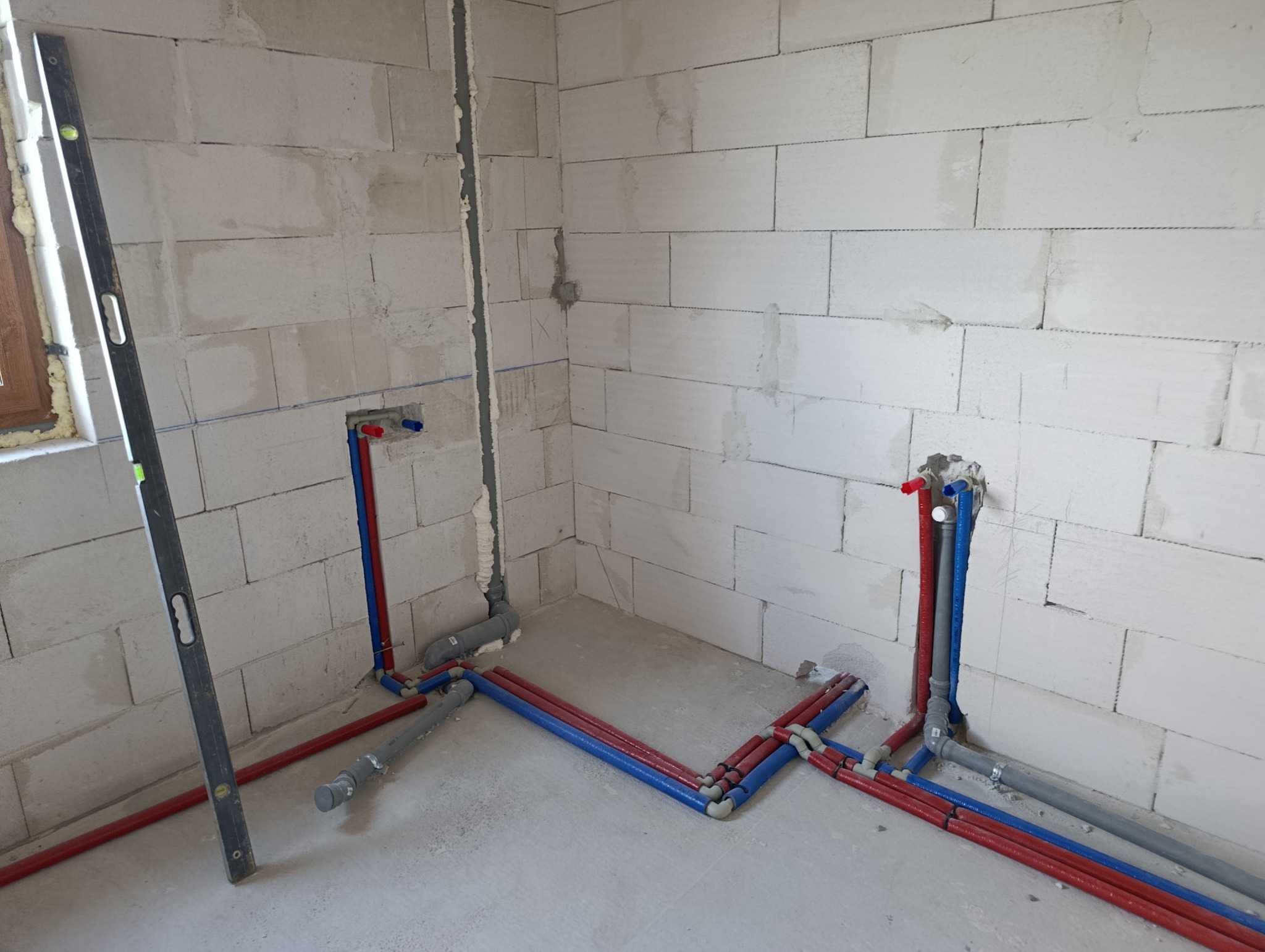 Wnętrze w trakcie prac hydraulicznych: czerwone i niebieskie rury instalacji wodnej ułożone na podłodze, pionowe podejścia w ścianach z betonu komórkowego, poziomica przy oknie.