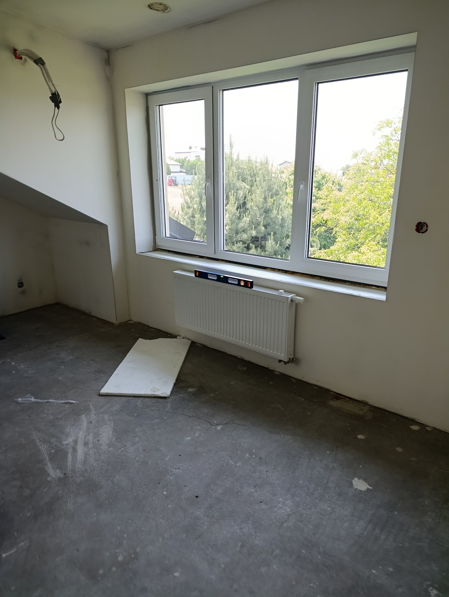 Biały grzejnik zamontowany pod oknem, sprawdzany poziomicą. W tle widok na zieleń. Na ścianie instalacja elektryczna. Surowa podłoga w szarym kolorze.