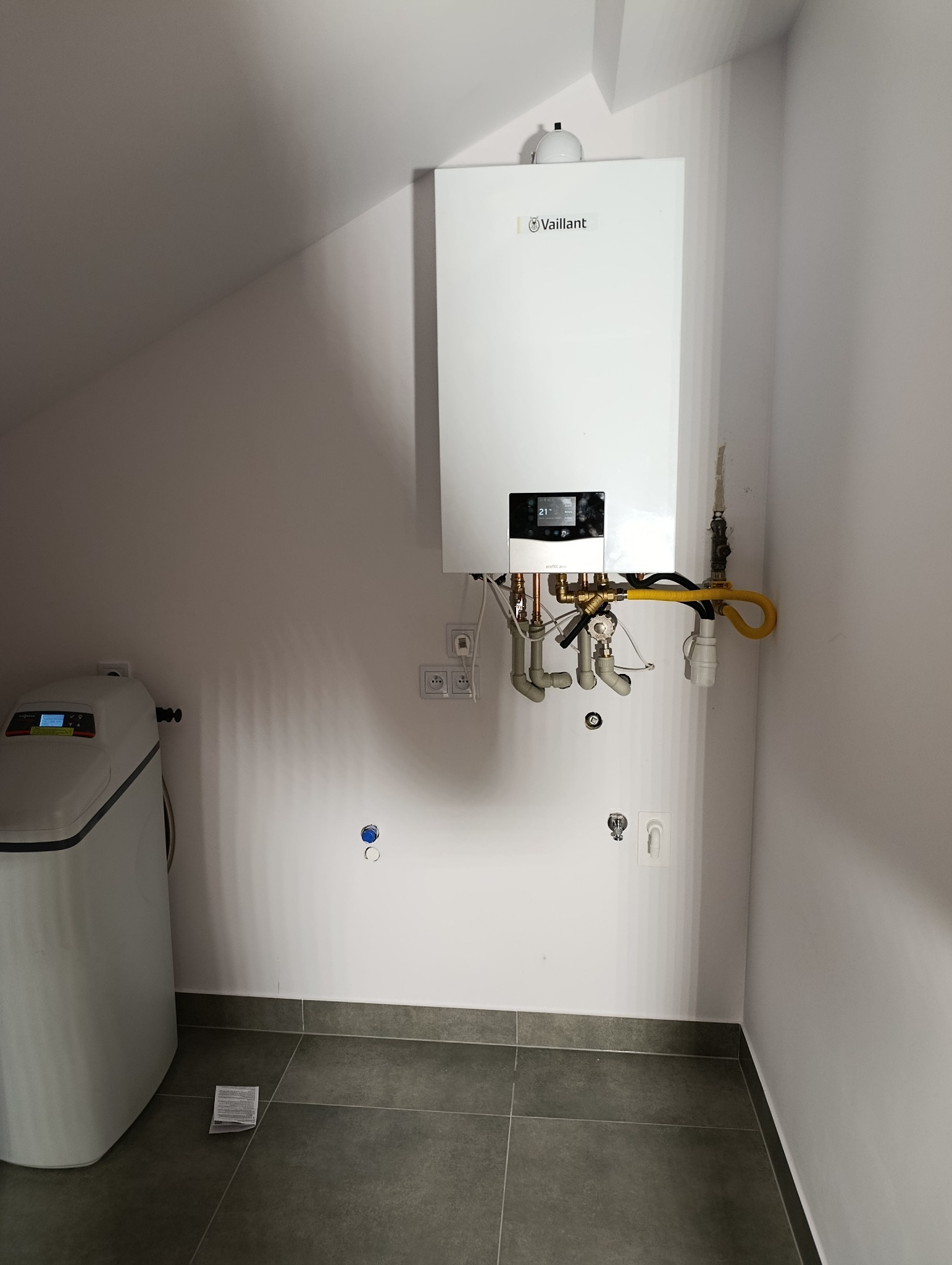 Biały piec gazowy Vaillant z żółtą rurą gazową na tle jasnych ścian i szarej podłogi. Widoczne przyłącza hydrauliczne i zmiękczacz wody po lewej.