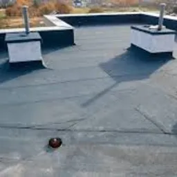 Czarny, płaski dach pokryty papą termozgrzewalną z kominkami wentylacyjnymi na tle jesiennego krajobrazu miejskiego, widziany z góry.