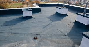 Czarny, płaski dach pokryty papą termozgrzewalną z kominkami wentylacyjnymi na tle jesiennego krajobrazu miejskiego, widziany z góry.