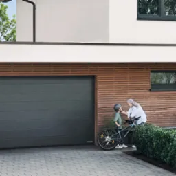 Nowoczesny dom z szarą bramą garażową segmentową i drzwiami wejściowymi. Babcia zapina kask wnukowi na rowerze. Drewniana elewacja i kostka brukowa.