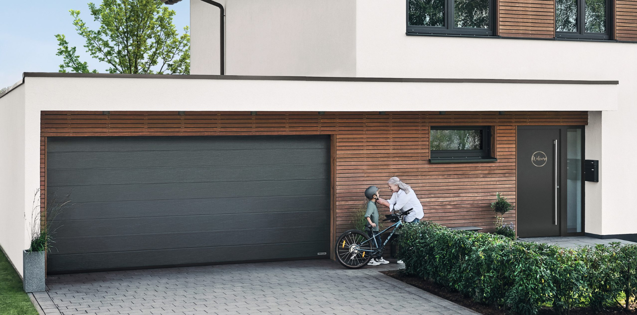 Nowoczesny dom z szarą bramą garażową segmentową i drzwiami wejściowymi. Babcia zapina kask wnukowi na rowerze. Drewniana elewacja i kostka brukowa.