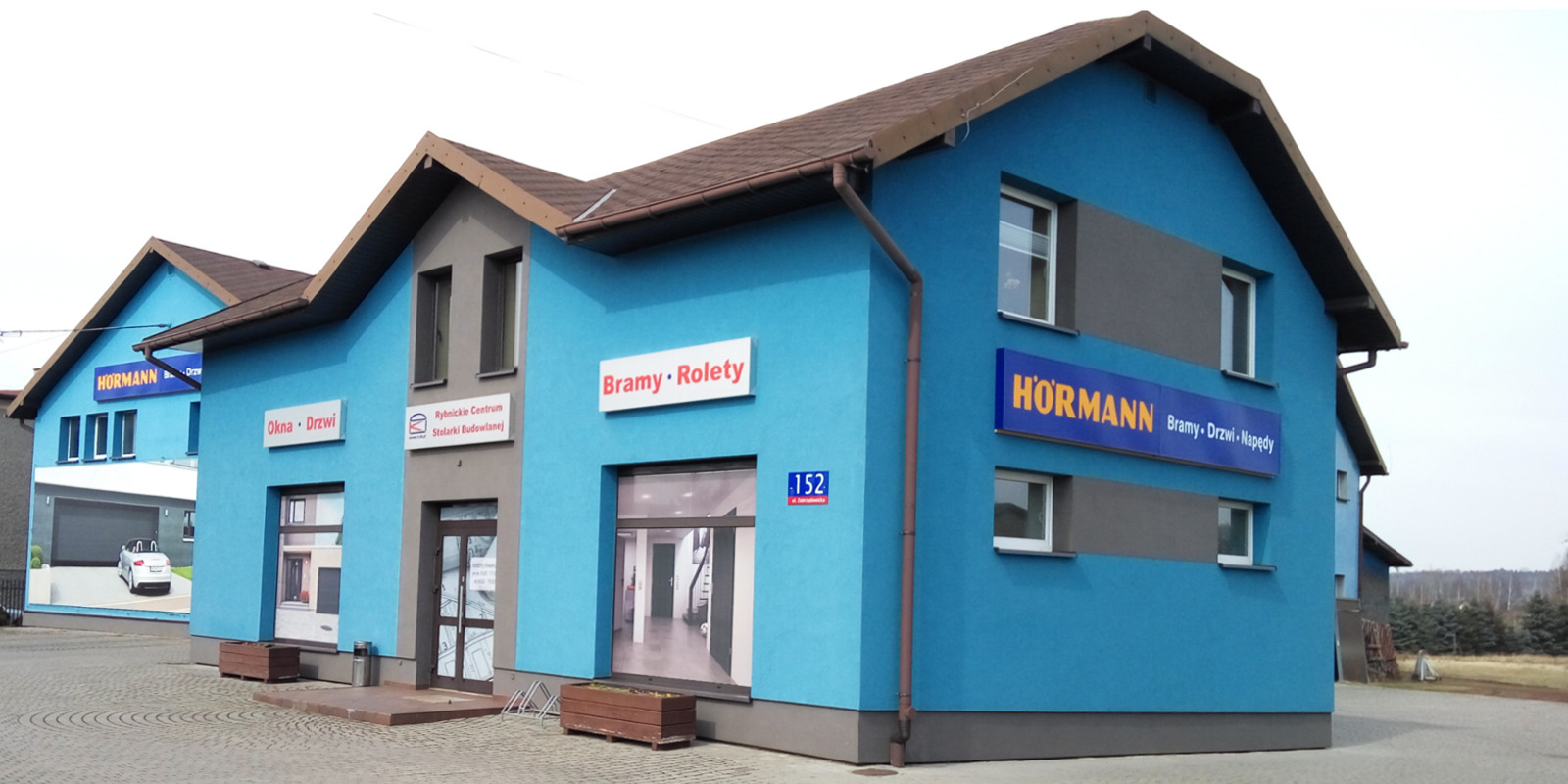 Niebieski budynek z szyldami reklamującymi okna, drzwi, bramy i rolety firmy Hormann oraz Rybnickie Centrum Stolarki Budowlanej. Widok na elewację z ulicy.