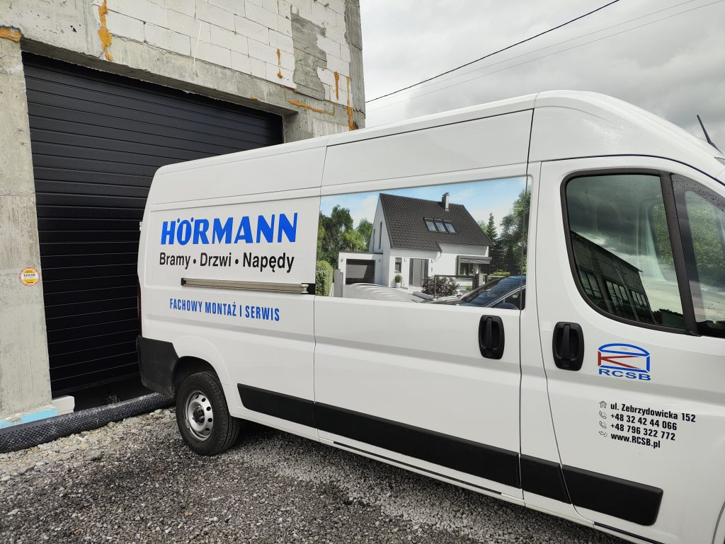 Czarna brama garażowa Hormann obok białego samochodu firmowego z reklamą bram, drzwi i napędów, na tle surowego budynku w trakcie budowy.