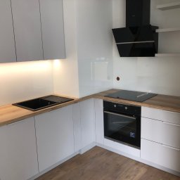 MK Meble Michał Kiszka - Minimalistyczna kuchnia z białymi frontami i drewnianym blatem. Czarny zlew i płyta indukcyjna. Nowoczesny okap nad płytą. Jasne, naturalne światło. Drewniana podłoga.