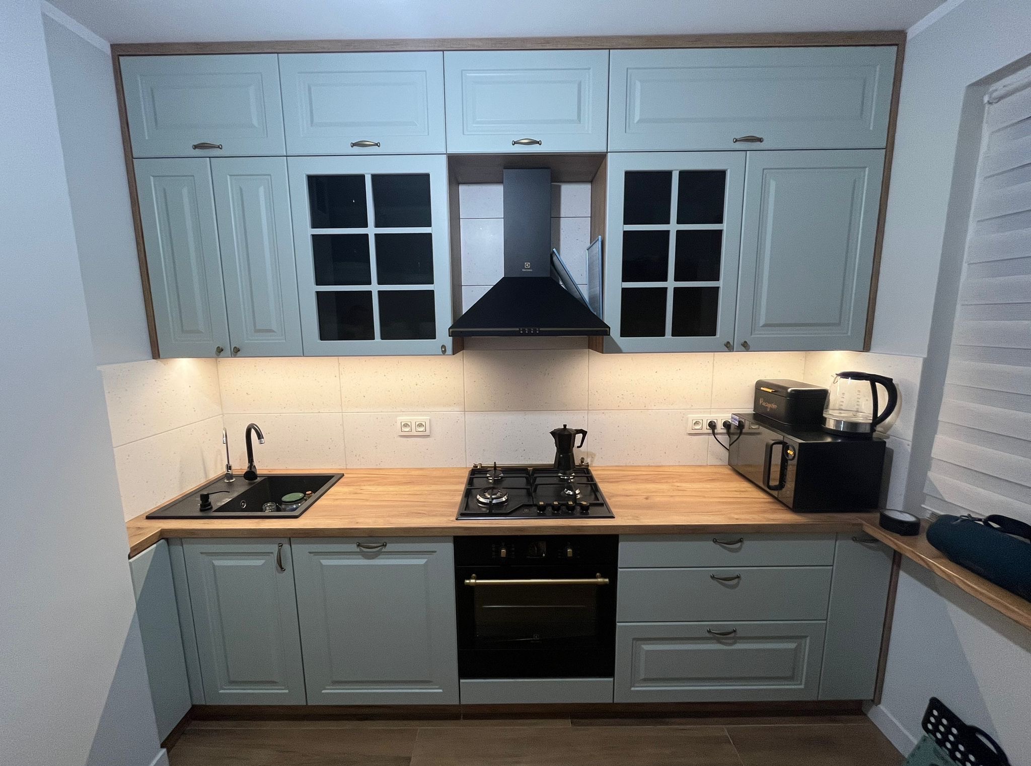 Jasnoszara kuchnia z drewnianym blatem i czarnymi sprzętami, w tym piekarnikiem i okapem. Widoczne górne szafki z przeszkleniami i ozdobnymi uchwytami.