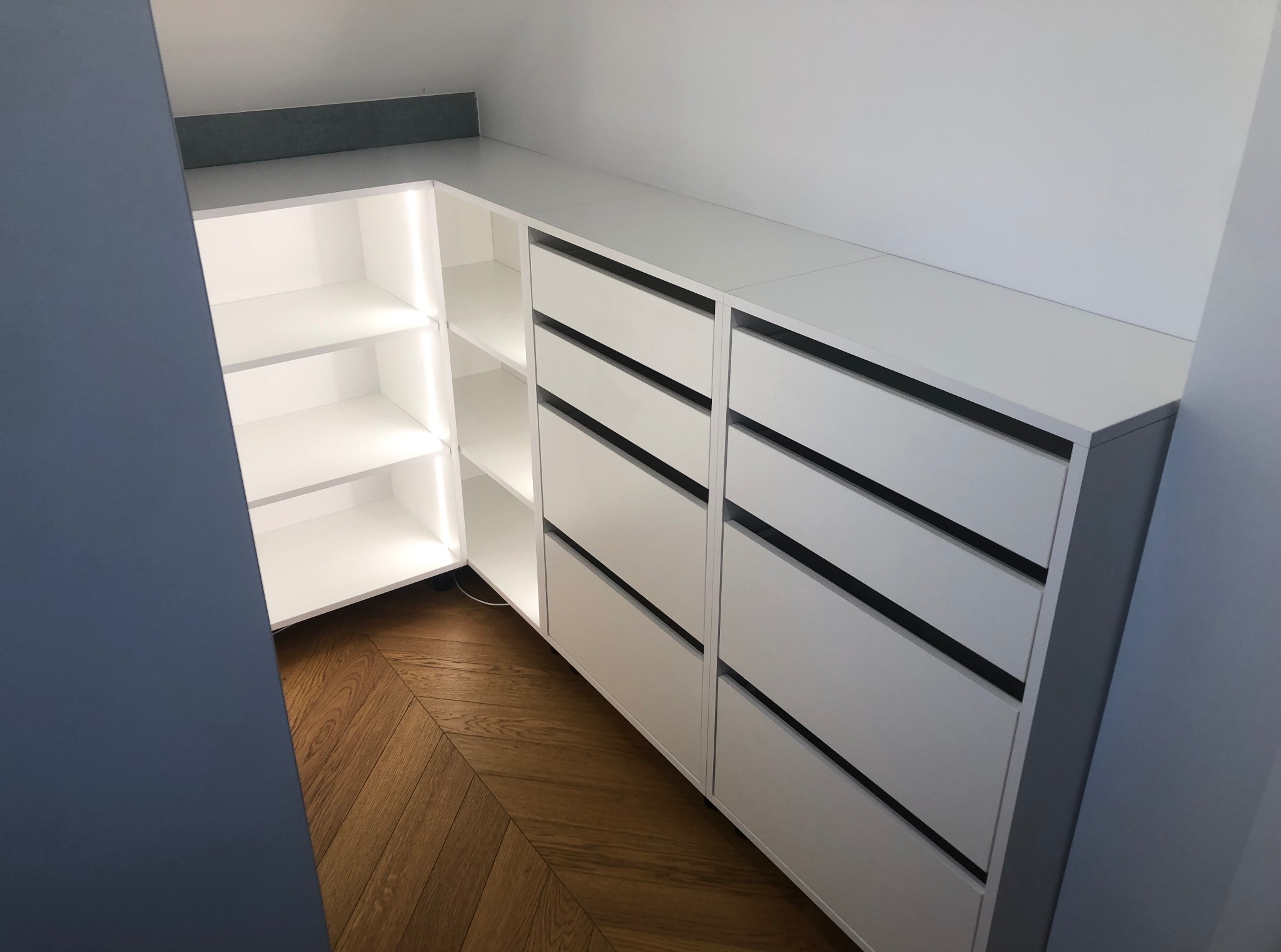 Biała szafa wnękowa z oświetleniem LED, z szufladami z czarnymi uchwytami, na parkiecie ułożonym w jodełkę. Nowoczesny design, wnętrze szafy z półkami.