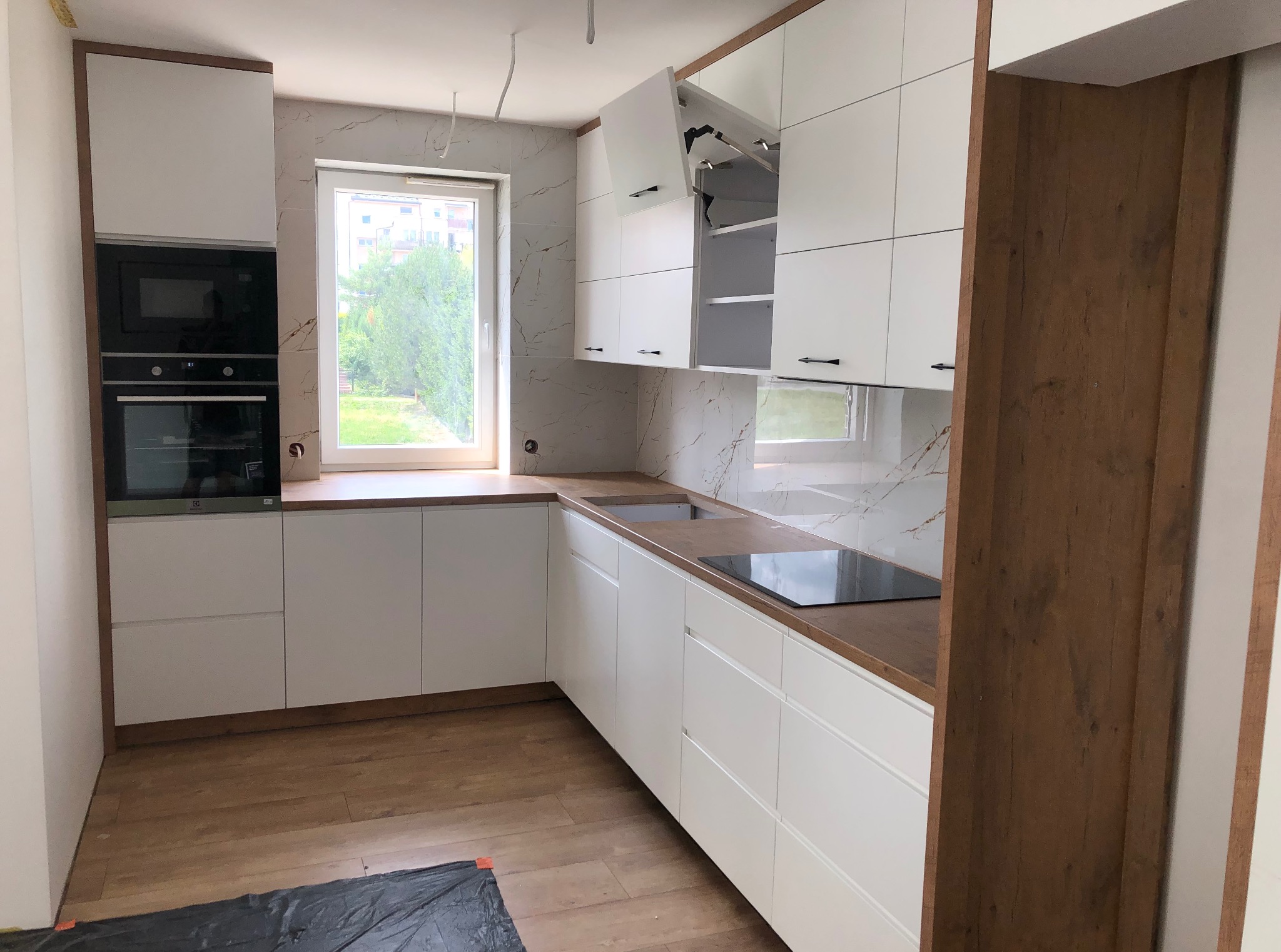 Nowoczesna, jasna kuchnia z białymi frontami i drewnianym blatem. Widoczne okno, marmurowe płytki i otwarte szafki górne z systemem podnoszenia.