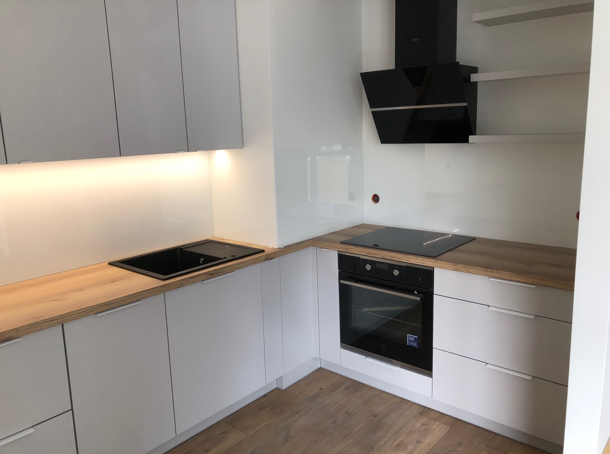 Minimalistyczna kuchnia z białymi frontami i drewnianym blatem. Czarny zlew i płyta indukcyjna. Nowoczesny okap nad płytą. Jasne, naturalne światło. Drewniana podłoga.