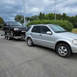 Srebrny Mercedes holuje na lawecie czarnego Range Rovera. Widoczny znak drogowy dla lawet. Transport pojazdów na tle zieleni i pochmurnego nieba.