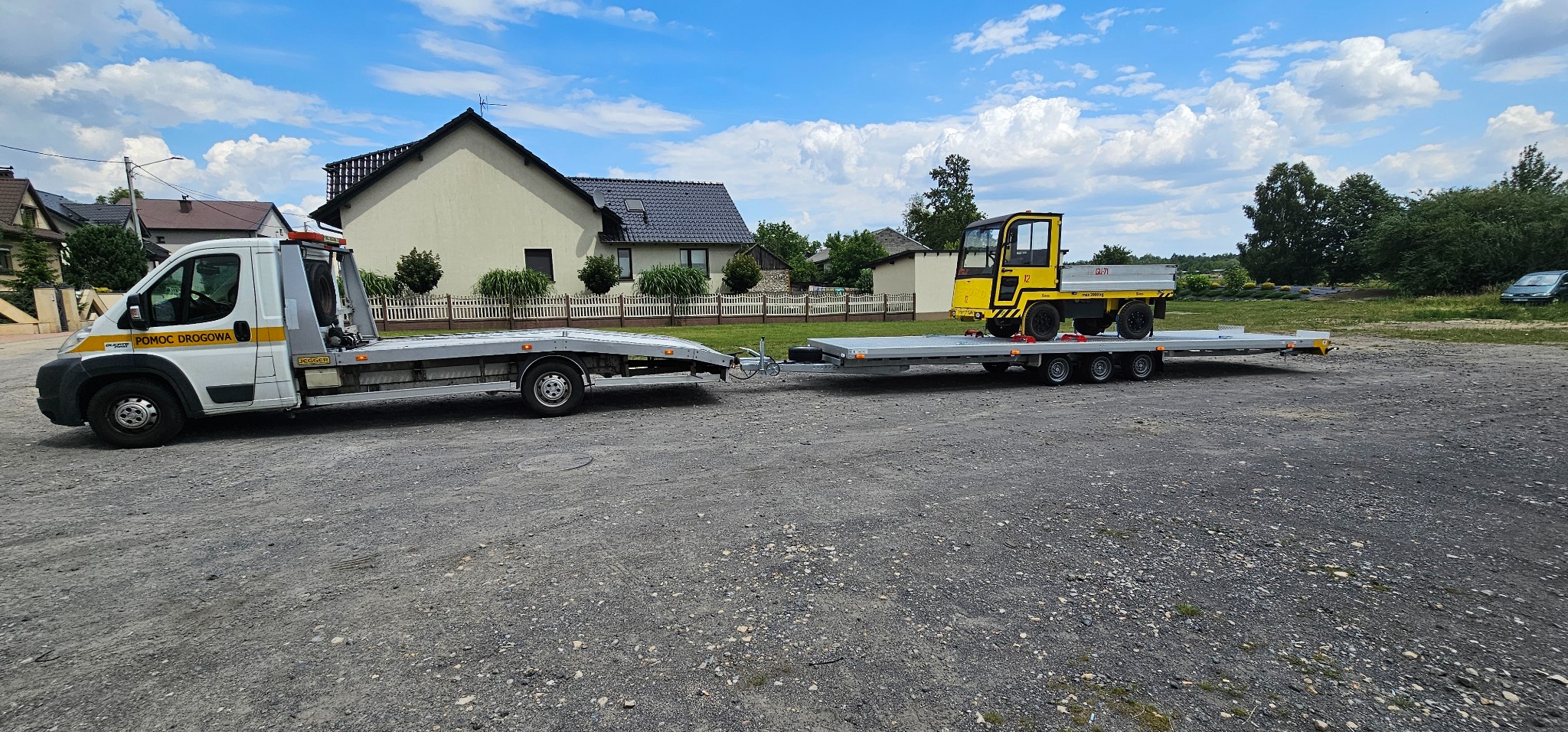 Laweta z żółtym pojazdem na platformie, zaparkowana na żwirowym terenie przed domem. Na lawecie widoczny mały pojazd roboczy.