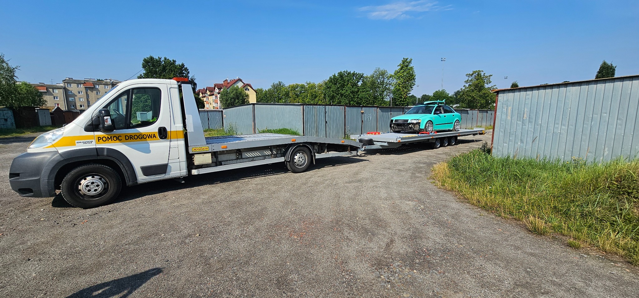 Laweta z turkusowym autem na platformie w Lubszy. Pojazd pomocy drogowej z żółtym pasem, widok z boku na tle zabudowań i zieleni.