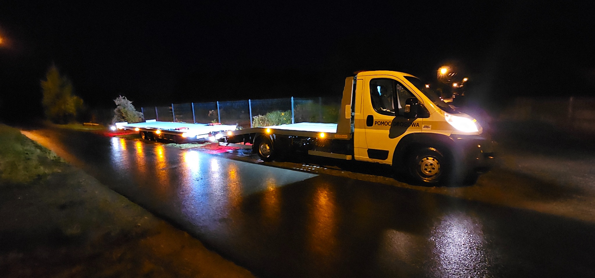 Żółta laweta 'Pomoc Drogowa' z przyczepą na mokrej, oświetlonej ulicy nocą. Widoczne światła lawety i odbicia w asfalcie. Przyczepa przewozi przewrócone drzewo.