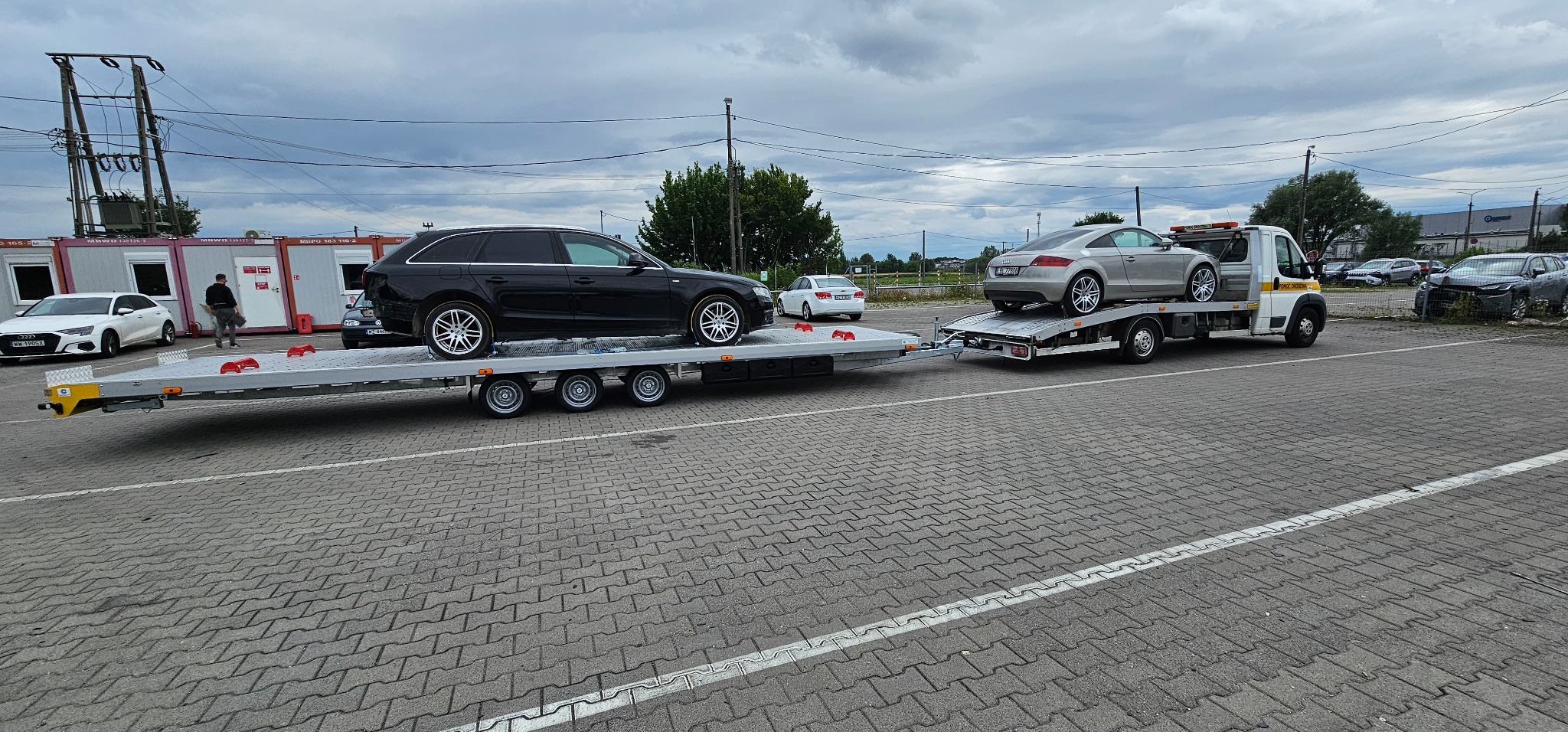 Laweta z dwoma autami: czarnym kombi Audi A4 i srebrnym Audi TT. Laweta stoi na parkingu, a w tle widać budynki kontenerowe i inne zaparkowane pojazdy.