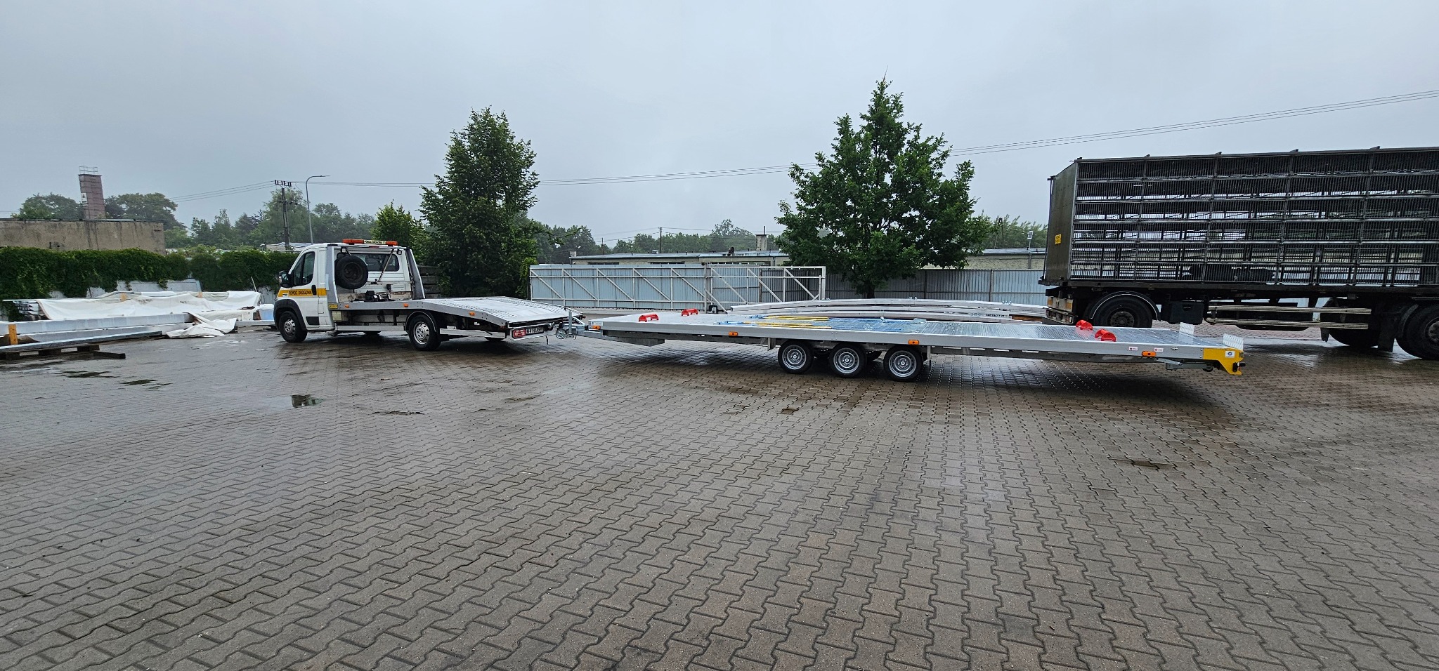 Laweta z platformą transportową na mokrym placu, widok z boku. Na platformie zabezpieczone ładunki, w tle zieleń i budynki. Pochmurny dzień.