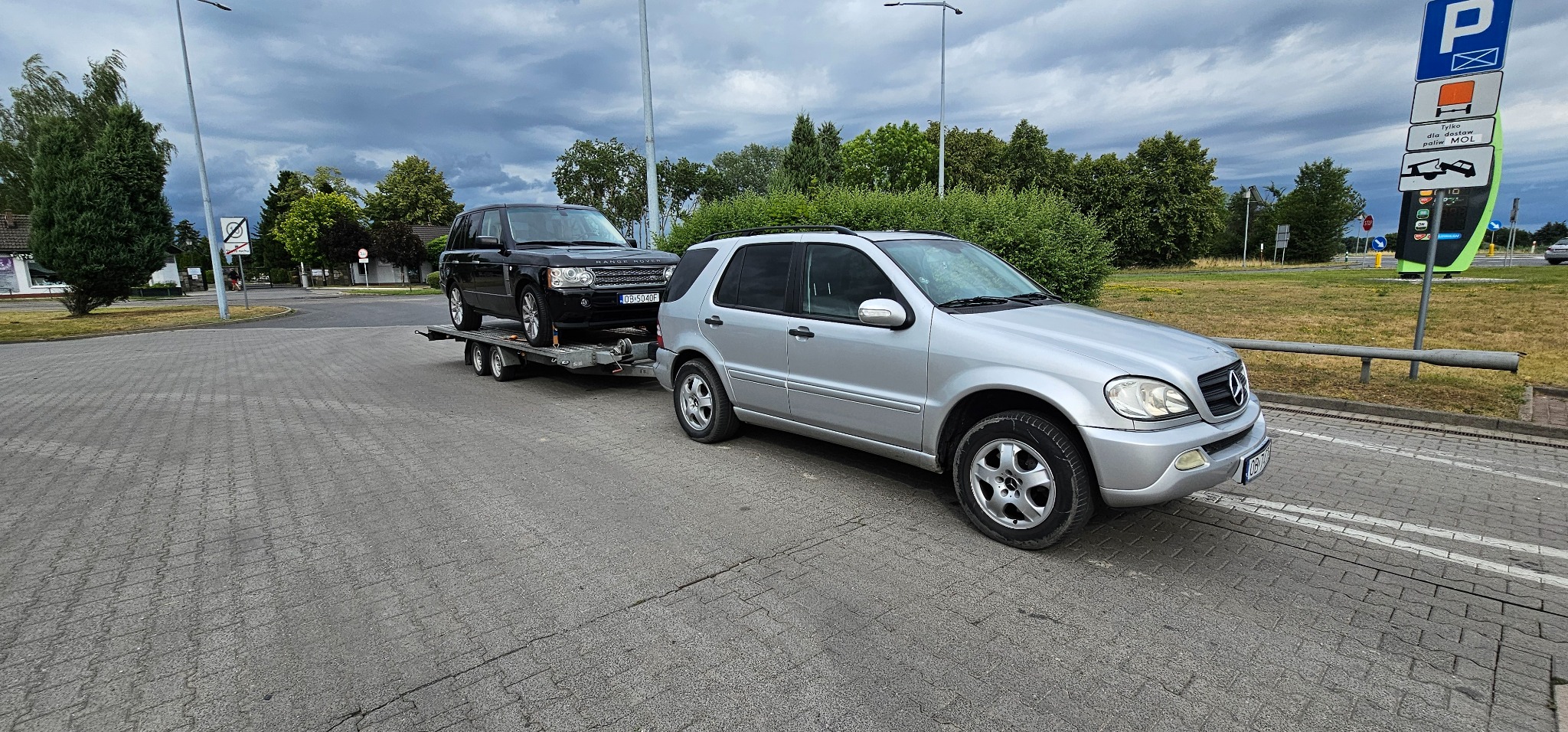 Srebrny Mercedes holuje na lawecie czarnego Range Rovera. Widoczny znak drogowy dla lawet. Transport pojazdów na tle zieleni i pochmurnego nieba.