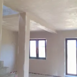 Wnętrze po nałożeniu tynku gipsowego w Jeleniej Górze, widok na ścianę, sufit i okno, z widocznymi śladami prac wykończeniowych.